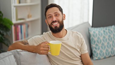 Genç İspanyol adam evdeki kanepede kahve içiyor.