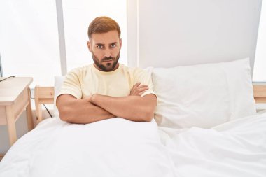 Young hispanic man sitting on bed with unhappy expression and arms crossed gesture at bedroom