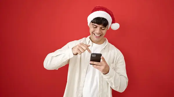 Junger Hispanischer Mann Mit Weihnachtsmütze Zeigt Auf Smartphone Vor Isoliertem — Stockfoto