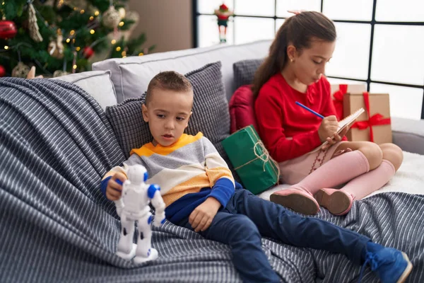 Brother and sister playing with robot and writing on notebook sitting on sofa by christmas tree at home