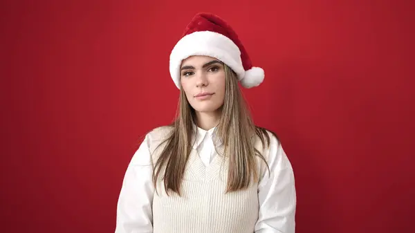 若いです美しいですヒスパニック女性立っています深刻な式身に着けていますクリスマス帽子以上隔離された赤の背景 — ストック写真