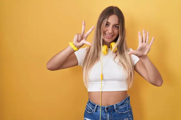 黄色い背景の上に立っている若い金髪の女性は 自信と幸せに微笑んでいる間 ヘッドフォンを示し 指番号8を指で指を指さす — ストック写真