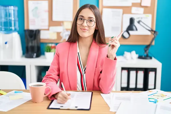 Young hispanic woman working at the office wearing glasses pointing with hand finger to the side showing advertisement, serious and calm face