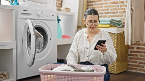 Young beautiful hispanic woman doing laundry using smartphone at laundry room