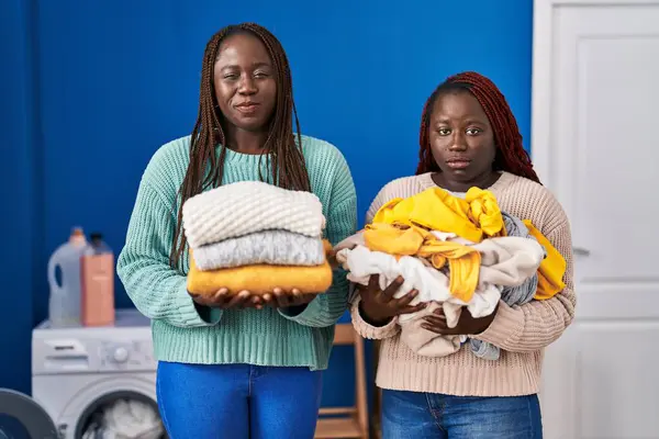Dwie Afrykańskie Kobiety Trzymające Złożone Pranie Prasowaniu Sceptyczne Nerwowe Marszczenie — Zdjęcie stockowe