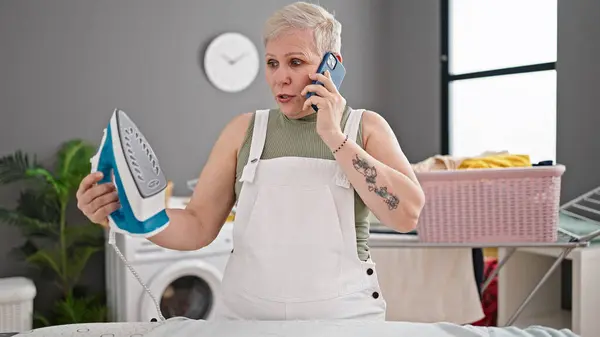Middle age grey-haired woman talking on smartphone holding ironing machine at laundry room