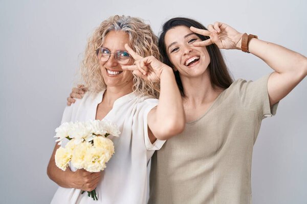Мать и дочь держат букет белых цветов, делая символ мира с пальцами на лице, улыбаясь весело показывая победу 