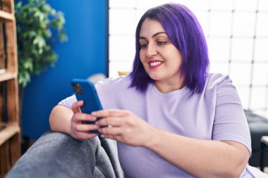 Genç, güzel, iri yarı bir kadın akıllı telefon kullanıyor. Evdeki koltukta oturuyor.