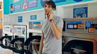 Genç İspanyol adam akıllı telefondan konuşuyor çamaşır makinesine yaslanıyor çamaşırhanede gülümsüyor.