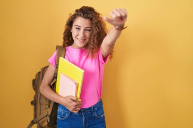 Öğrenci sırt çantası takan, elinde kitaplarla olumlu jestler yapan, gülümseyen ve başarı için mutlu genç beyaz bir kadın. kazanan jest. 