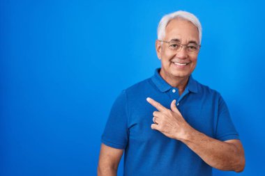 Mavi arka planda duran gri saçlı orta yaşlı bir adam. Yüzünde mutlu ve doğal bir ifadeyle elleri ve parmaklarıyla gülümseyen bir gülümseme. 