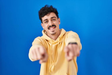 Mavi arka planda duran İspanyol adam sana ve kameraya parmaklarıyla bakıyor, gülümsüyor, pozitif ve neşeli. 