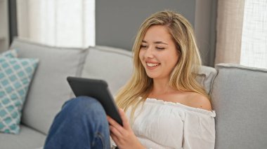 Young blonde woman using touchpad sitting on sofa at home