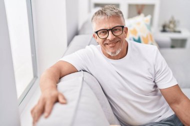 Orta yaşlı, kır saçlı, gülümseyen bir adam. Evdeki kanepede oturuyor.