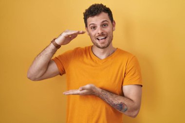 Young hispanic man standing over yellow background gesturing with hands showing big and large size sign, measure symbol. smiling looking at the camera. measuring concept. 