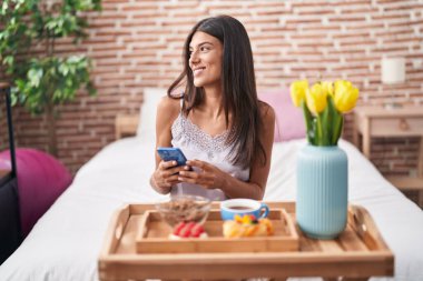 Young beautiful hispanic woman having gift breakfast using smartphone at bedroom