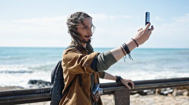 Sırt çantası takan genç İspanyol turist deniz kenarında akıllı telefondan selfie çekiyor.