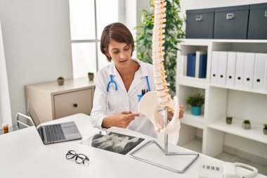 Young beautiful hispanic woman doctor pointing with finger to anatomical model of vertebral column at clinic