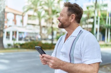 Orta yaşlı bir adam sokakta akıllı telefon kullanarak kendinden emin gülümsüyor.