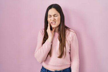 Young brunette woman standing over pink background touching mouth with hand with painful expression because of toothache or dental illness on teeth. dentist 