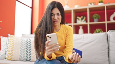 Genç, İspanyol bir kadın akıllı telefon ve kredi kartıyla alışveriş yapıyor. Evdeki koltukta oturuyor.