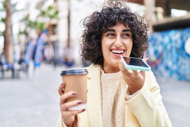 Orta Doğulu genç bir kadın akıllı telefonla konuşuyor sokakta kahve içiyor.