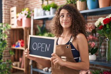 Kıvırcık saçlı İspanyol kadın çiçekçide çalışıyor açık tabela tutuyor, gülümsüyor ve başka tarafa bakıyor.. 