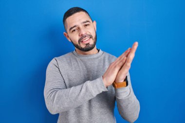 Hispanic man standing over blue background clapping and applauding happy and joyful, smiling proud hands together 