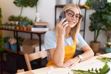 Young blonde woman florist smiling confident talking on smartphone at flower shop
