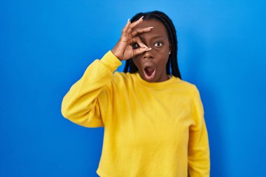 Beautiful black woman standing over blue background doing ok gesture shocked with surprised face, eye looking through fingers. unbelieving expression. 