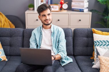 Genç İspanyol adam evdeki koltukta dizüstü bilgisayar kullanıyor.
