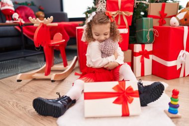 Sevimli sarışın kız oyuncak ayı hediyesini açıyor. Evde Noel ağacının yanında oturuyor.