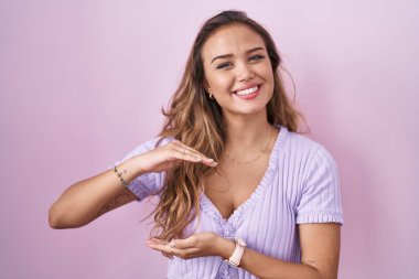 Young hispanic woman standing over pink background gesturing with hands showing big and large size sign, measure symbol. smiling looking at the camera. measuring concept. 