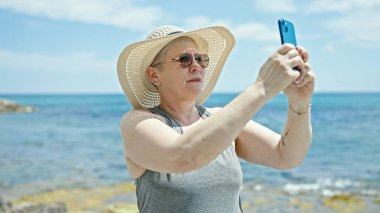 Orta yaşlı, kır saçlı bir turist kumsalda akıllı telefondan fotoğraf çekiyor.