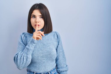 Mavi arka planda dikilen genç İspanyol kadın, parmakları dudaklarında sessiz olmak istiyor. sessizlik ve gizli konsept. 