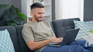 Genç İspanyol adam evdeki koltukta dizüstü bilgisayar kullanıyor.