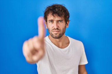 Mavi arka planda dikilen İspanyol genç adam parmağını kaldırmış ve kızgın bir ifadeyle işaret ediyor, hiçbir hareket göstermiyor. 