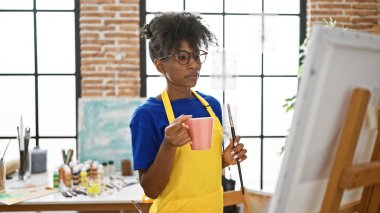Afrikalı Amerikalı kadın ressam sanat stüdyosunda elinde bir fincan kahve tutuyor.
