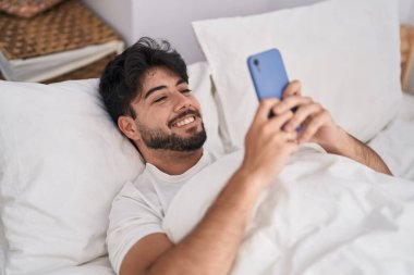 Genç İspanyol adam yatakta akıllı telefon kullanıyor.