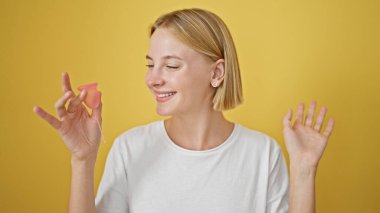 Genç sarışın kadın gülümsüyor. Aybaşı bardağını izole edilmiş sarı arka planda tutuyor.