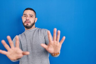 Hispanic man standing over blue background afraid and terrified with fear expression stop gesture with hands, shouting in shock. panic concept. 