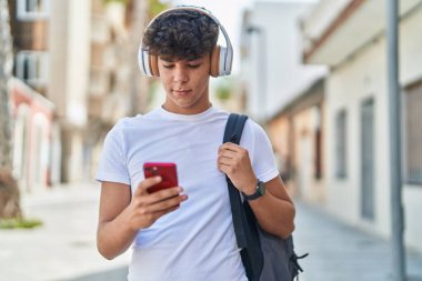 Young hispanic teenager student listening to music with relaxed expression at street