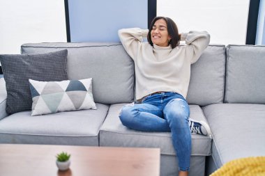 Young hispanic woman relaxed with hands on head sitting on sofa at home