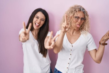 Anne ve kızı pembe arka planda yan yana durur ve iki elin parmaklarını göstererek gülümseyerek zafer işareti yaparlar. İki numara.. 