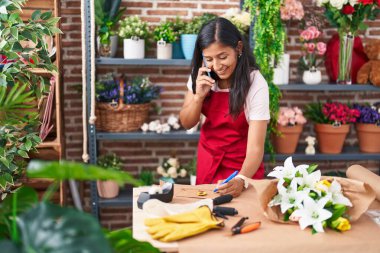 Young beautiful hispanic woman florist talking on smartphone writing on envelope letter at flower shop