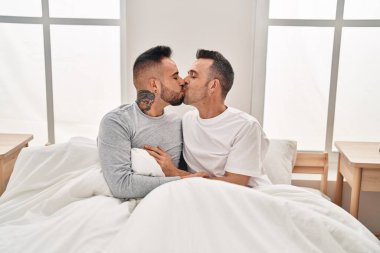 İki erkek yatak odasında birbirlerine sarılıp öpüşüyorlar.