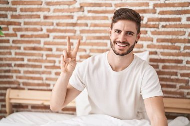 Beyaz bir adam zafer jesti yapıyor. Yatak odasında oturuyor.