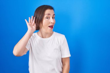 Middle age hispanic woman standing over blue background smiling with hand over ear listening an hearing to rumor or gossip. deafness concept. 