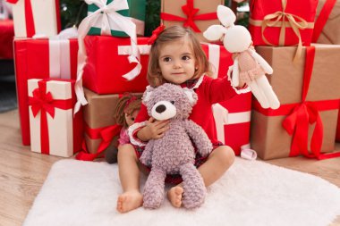 Sevimli sarışın bebek elinde oyuncak ayıyla evde Noel hediyeleriyle oturuyor.