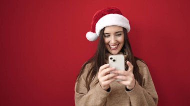 Güzel İspanyol kadın, yılbaşı şapkası takıyor ve izole edilmiş kırmızı arka plan üzerinde akıllı telefon kullanıyor.